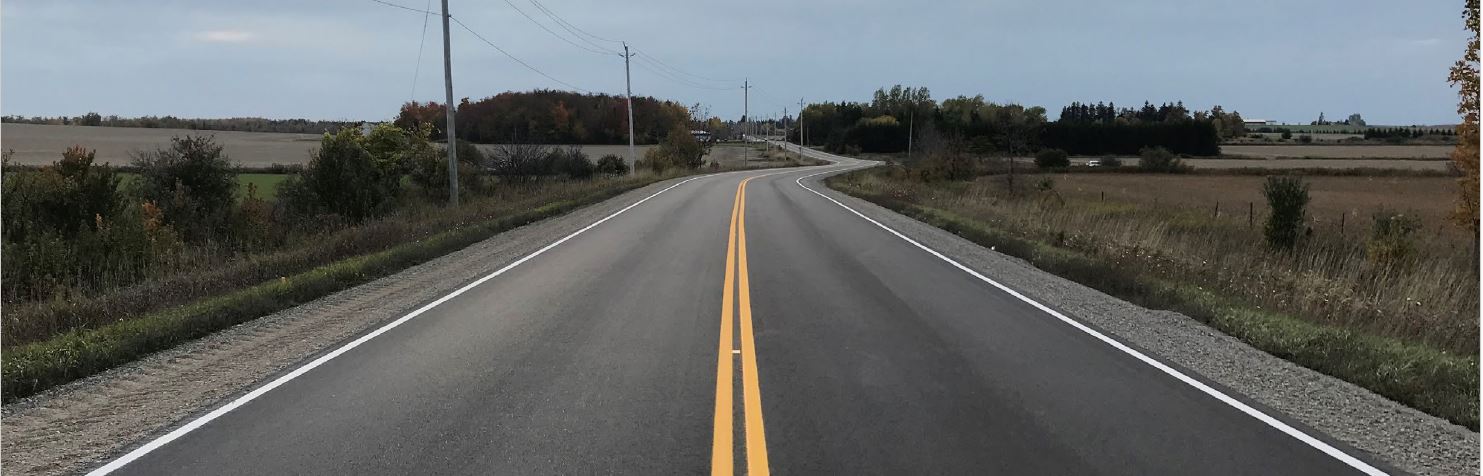 A paved road in a rural area winds through a field with hydro polls along side it.