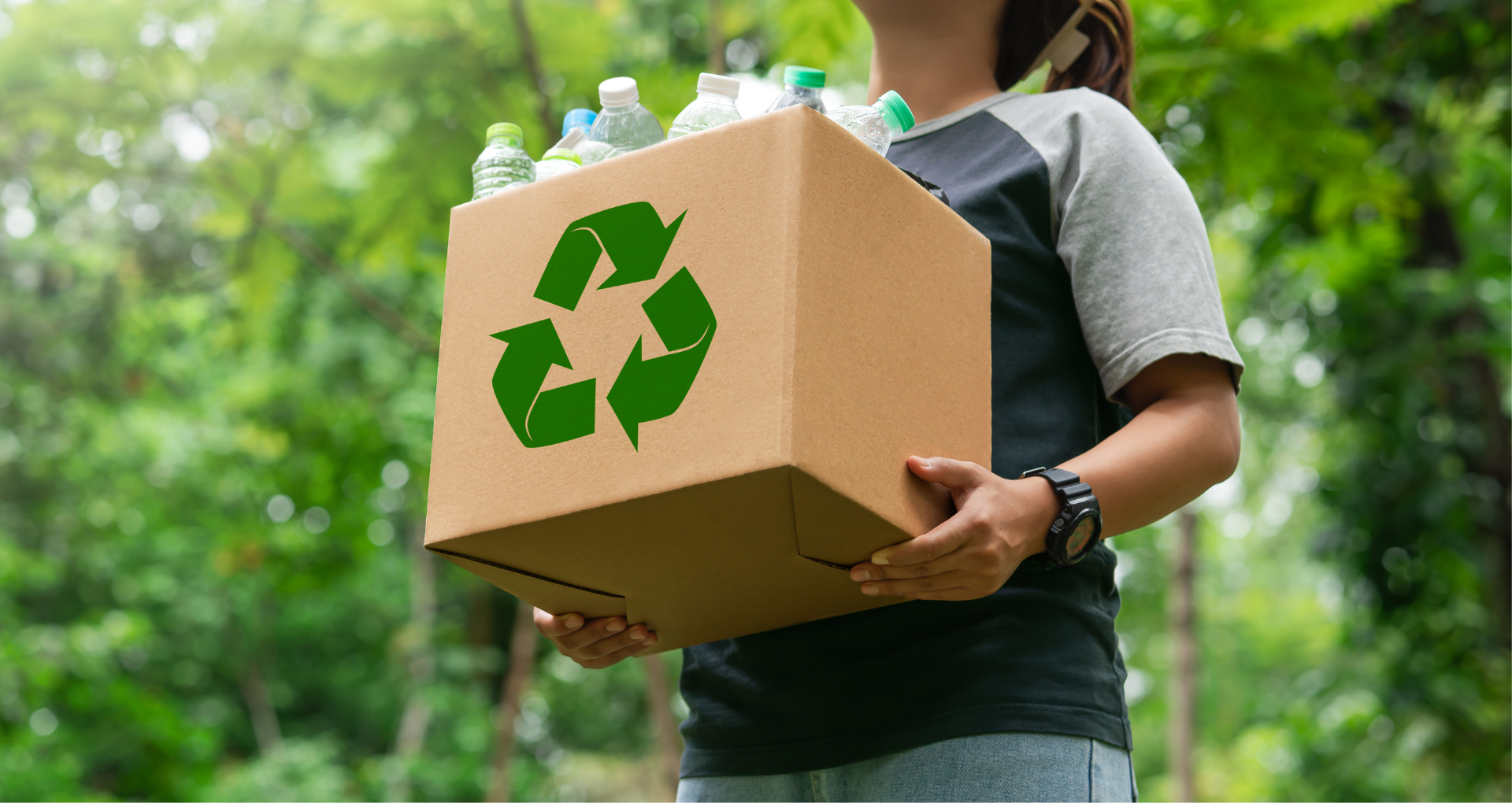 A person stands in front of green trees, holding a cardboard box with a green recycling symbol on it