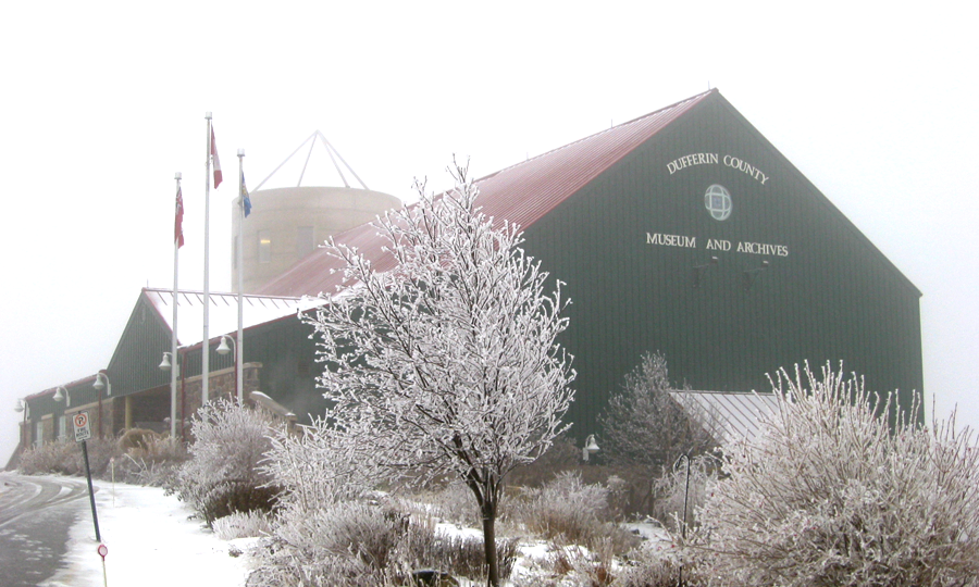 The Museum of Dufferin, a large green building designed to look like a barn, surrounded by icy trees and snow on a winter day.